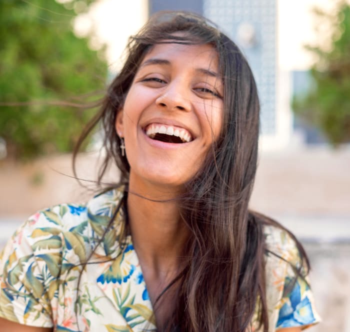 Woman with long dark hair and a colourful shirt sits outside and laughs while looking at the camera.