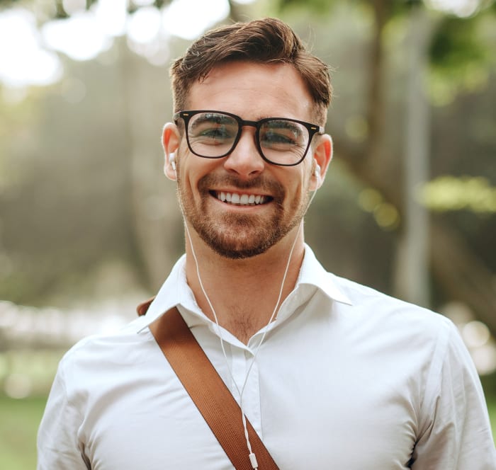 Young man with glasses stands outside wearing a messenger bag over his shoulder and has headphones in. He is smiling at the camera.