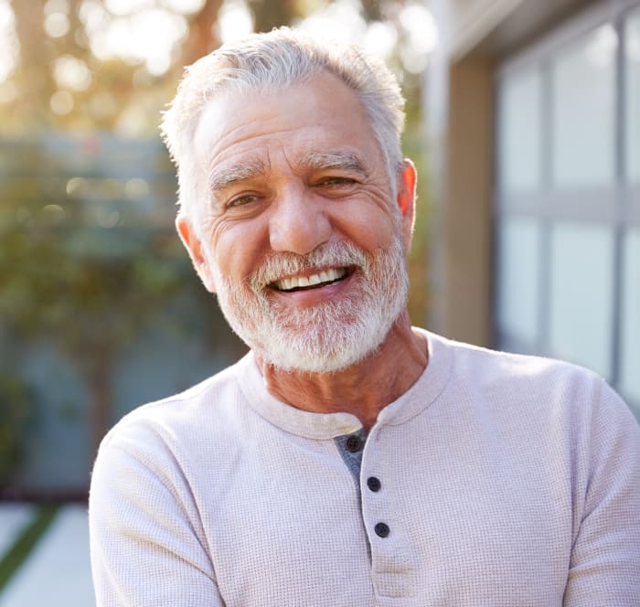 An older man with short white hair and bread smiles at the camera while standing outside.
