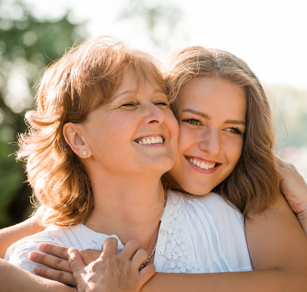 A woman with short hair is sitting outside being hugged from behind by a younger woman with longer hair. They are both smiling.