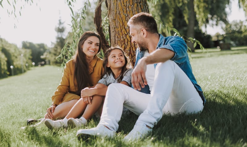 A young family with a little girl sits outside near a tree on a sunny day. They are all looking at eachother smiling.