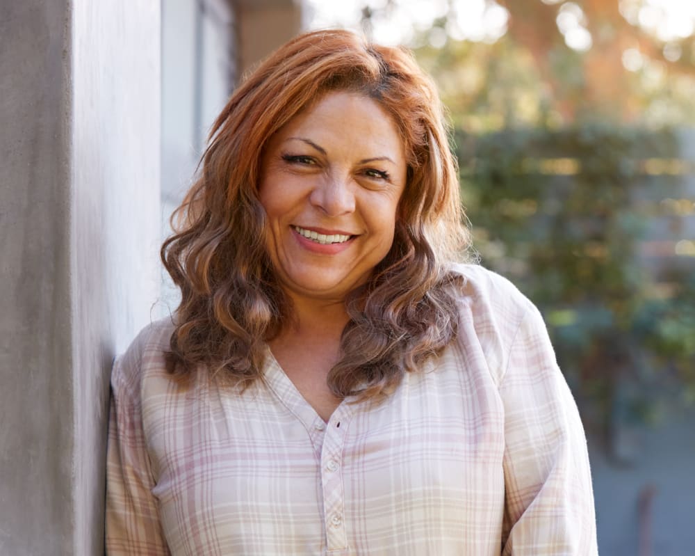A woman with long hair stands outside against a wall on a sunny day and is smiling at the camera.