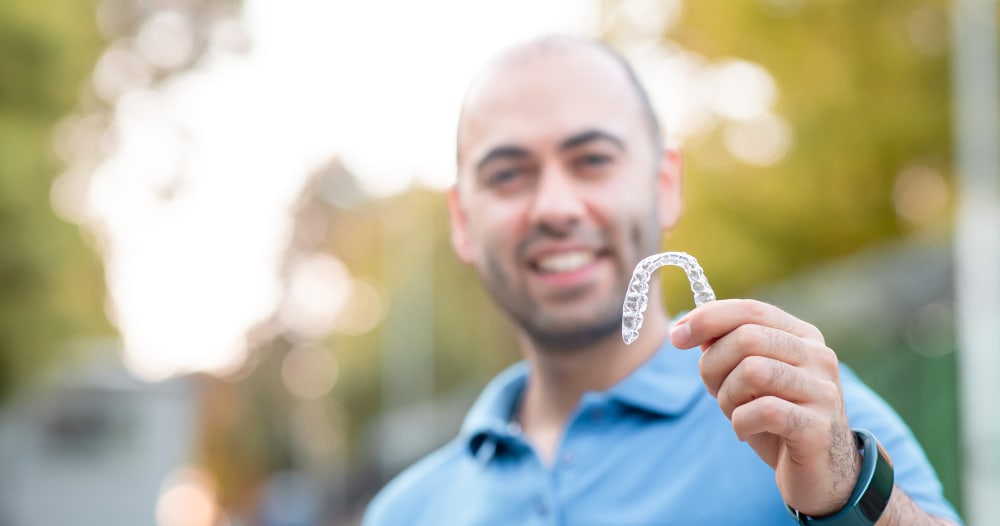 A man wearing a blue shirt stands outside on a nice day. He is just out of focus while holding up a pair of clear aligners which are in focus.