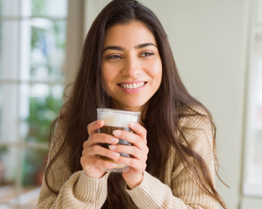 A young woman with long dark hair sits inside smilng while looking off to the side. She is holding a cup of coffee in front of her with both hands.